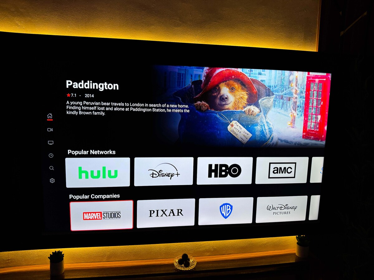 A large smart TV displays an animated movie selection screen with various streaming services, including Hulu and Disney+. The TV is positioned on a sleek console, illuminated by soft ambient lighting that enhances the viewing experience.