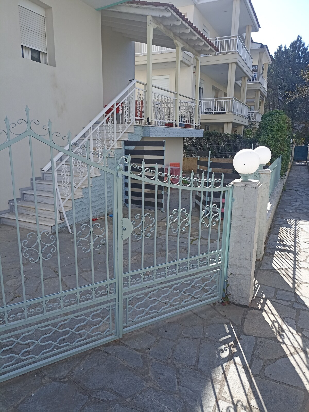 An ornate metal gate is showcased at the entrance, leading to a stone pathway. The property features a light-colored building with multiple balconies. Lush greenery surrounds the area, adding a touch of nature. Soft sunlight creates gentle shadows on the ground.