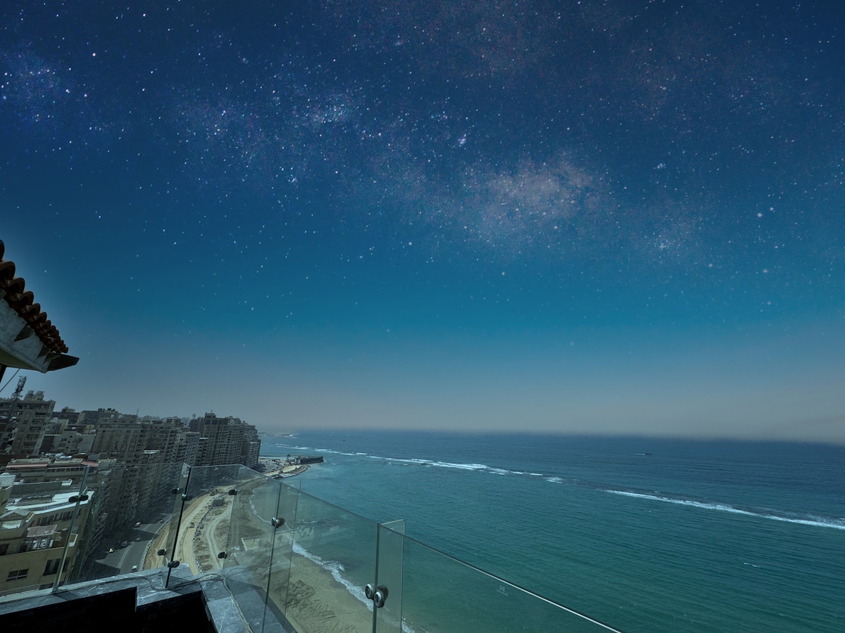 A breathtaking view of the Mediterranean Sea is captured from an elevated terrace, showcasing the coastline and gentle waves under a starlit sky. City buildings line the shore, and a clear horizon beckons from beyond the water.