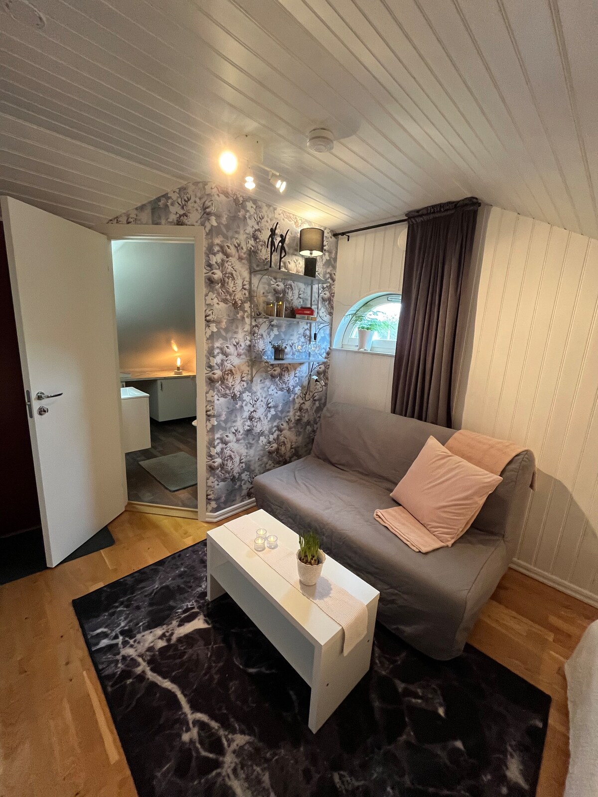 A cozy sitting area features a soft sofa adorned with a light pillow, placed beside a small, minimalist coffee table. The walls display a floral design, and a curved window allows natural light to brighten the space. A door leads to an unseen area.