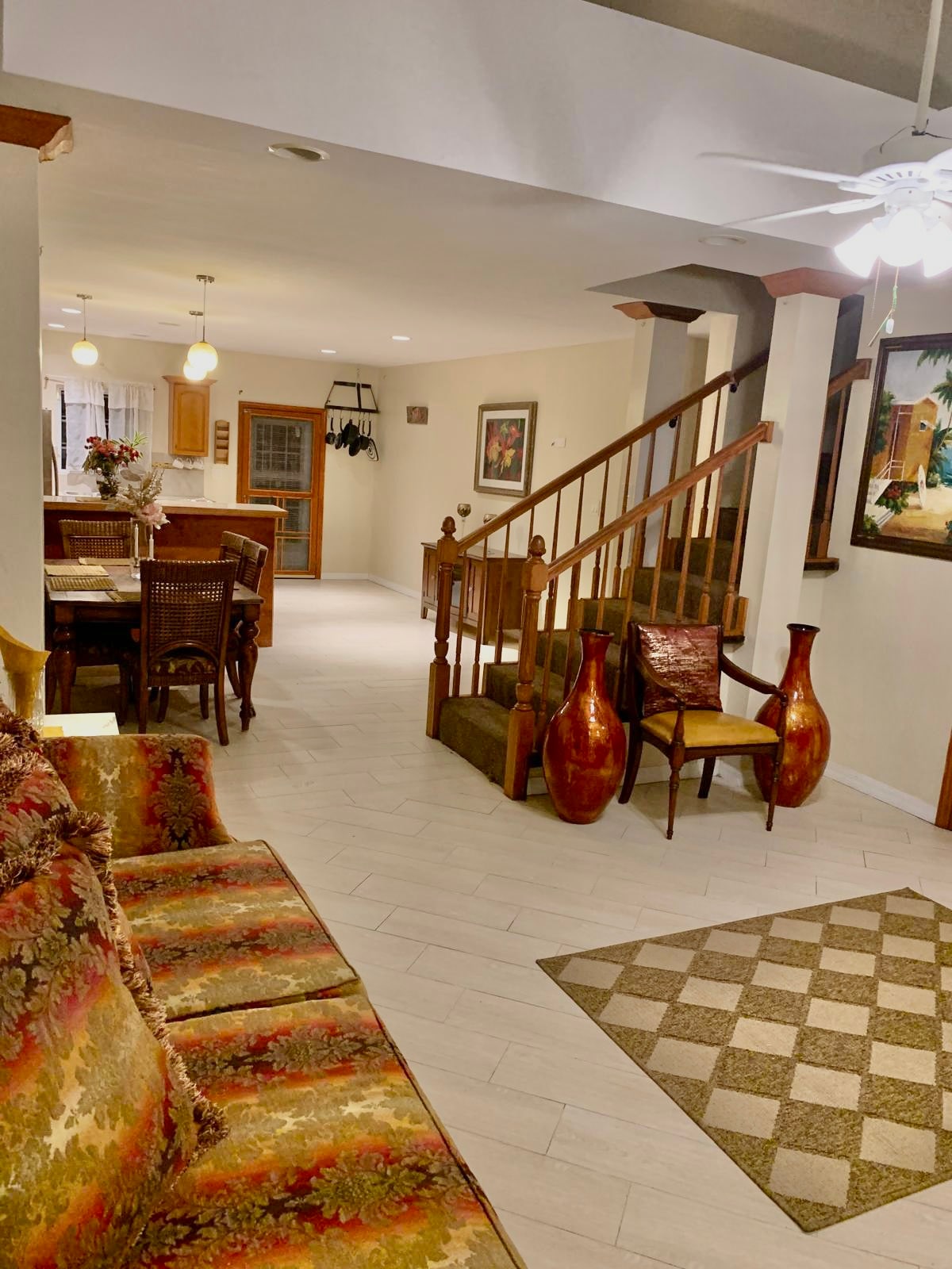 A spacious living area features a patterned area rug and a colorful sofa. The room is enhanced by a dining area visible in the background, along with a staircase leading to the upper level. Natural light is allowed in through multiple windows.