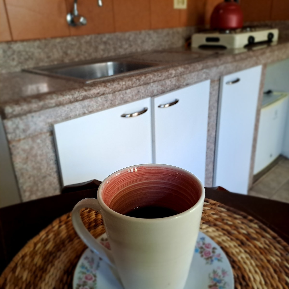 A ceramic mug filled with dark beverage is placed on a decorative plate. In the background, a kitchen features granite countertops, a sink, and a stove with a kettle atop. Storage cabinets are visible beneath the counter.