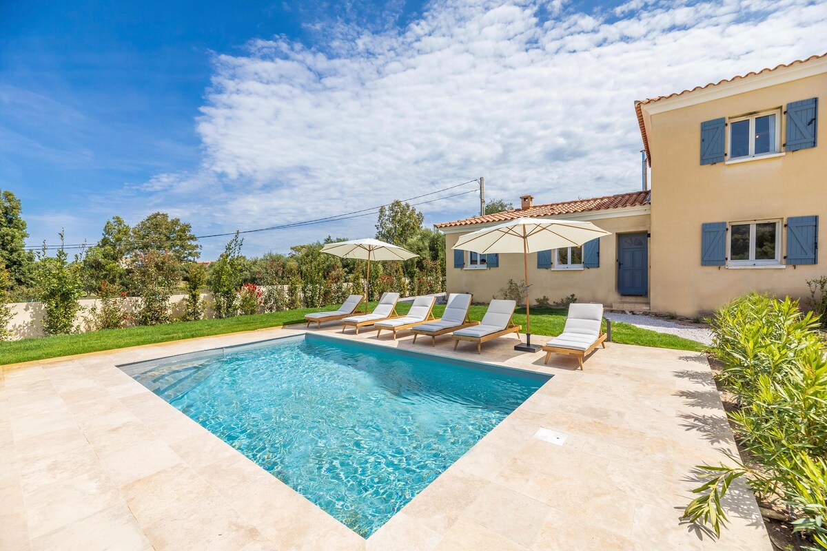 A private pool is framed by a spacious terrace featuring several lounge chairs under large umbrellas. Lush greenery surrounds the area, with a charming villa visible in the background, showcasing blue shutters and a warm exterior.