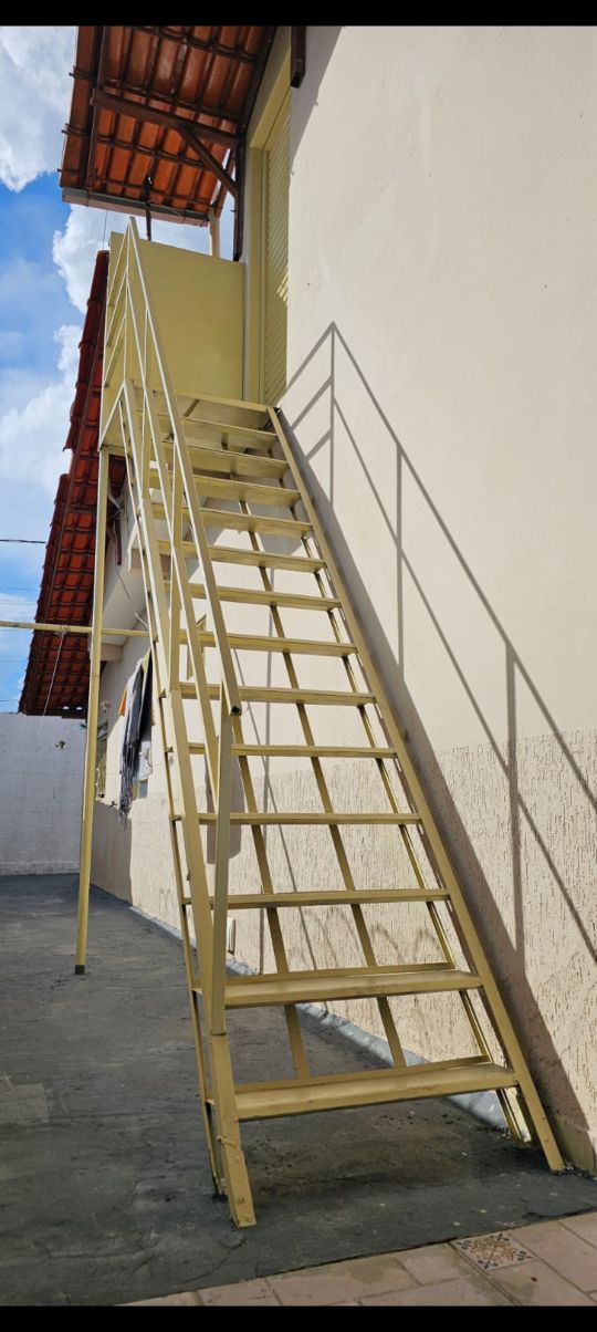 An exterior view of a yellow staircase leading up to a second level, positioned against a light-colored wall. The staircase features a railing and a clear pathway, with a partially cloudy sky visible above.