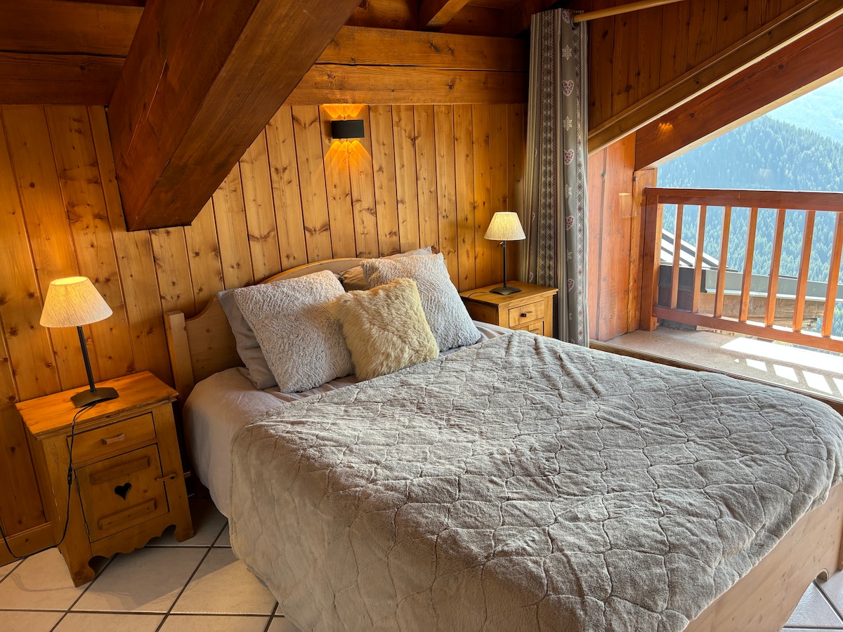 A cozy bedroom features a spacious bed with soft bedding and decorative pillows. Bedside tables hold lamps that create a warm glow. Wood paneling provides a rustic touch, while large windows frame mountain views and allow natural light to fill the room.