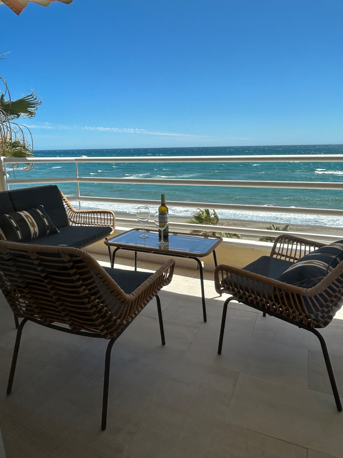 A small outdoor seating area is presented, featuring two rattan chairs and a glass table. The ocean view is unobstructed, with waves visible against a clear blue sky. The atmosphere appears inviting for relaxation or enjoying meals while overlooking the beach.
