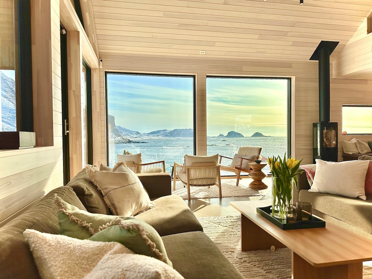 Spacious living area features large windows framing stunning sea views. A comfortable sofa is adorned with soft cushions, while a wooden coffee table holds a small vase of flowers. Two chairs are positioned nearby, inviting relaxation and enjoyment of the scenic landscape.