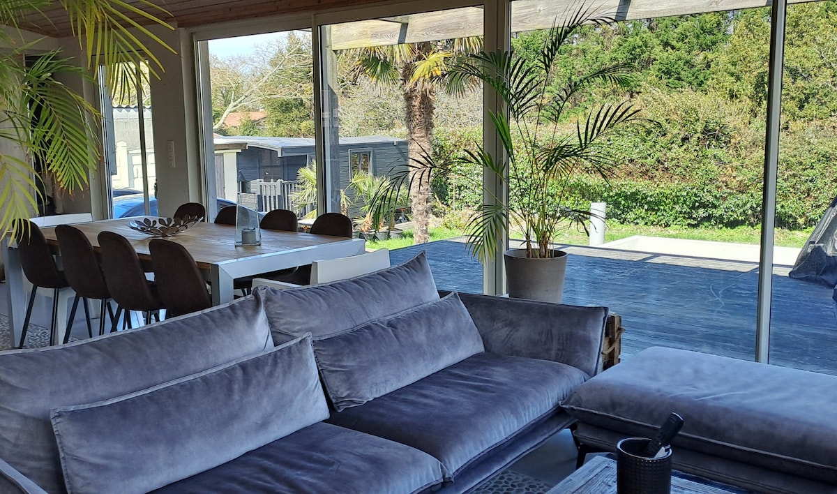 A spacious living area features large glass doors that provide a view of the outdoor terrace and garden. A comfortable gray sofa is positioned near a coffee table, complemented by stylish dining chairs at a sleek dining table in the background. Lush plants add a touch of greenery.