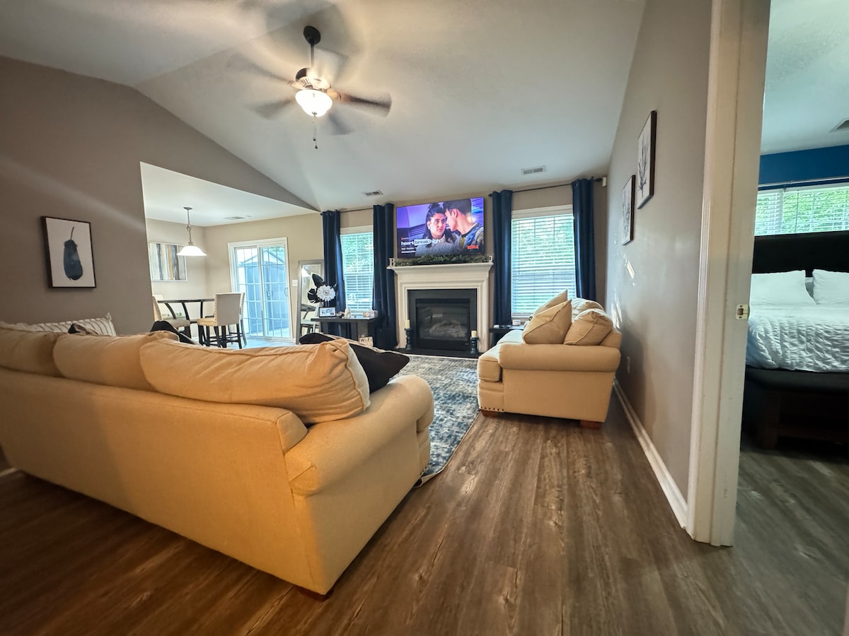 A spacious living area features plush sofas positioned around a cozy fireplace. A mounted television is visible above the mantle, providing entertainment. Natural light streams in through large windows, enhancing the inviting atmosphere. The room connects to a dining area visible in the background.