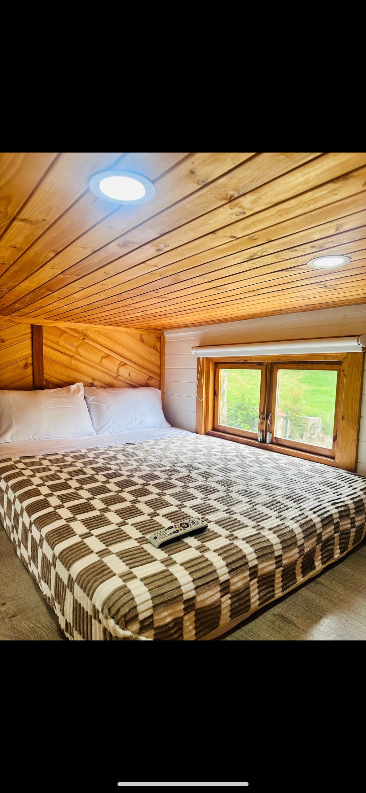A cozy sleeping area features a spacious bed with a checkered patterned mattress. Two large windows allow natural light to fill the room, framed by wooden accents and inviting views of the surrounding nature.