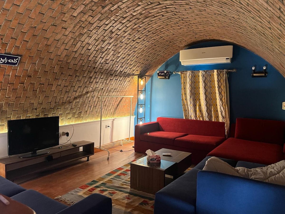 An inviting living area features a curved brick ceiling and a mix of red and blue sofas arranged around a low coffee table. A colorful area rug lies beneath, while soft lighting highlights the walls. An air conditioning unit ensures comfort in the space.