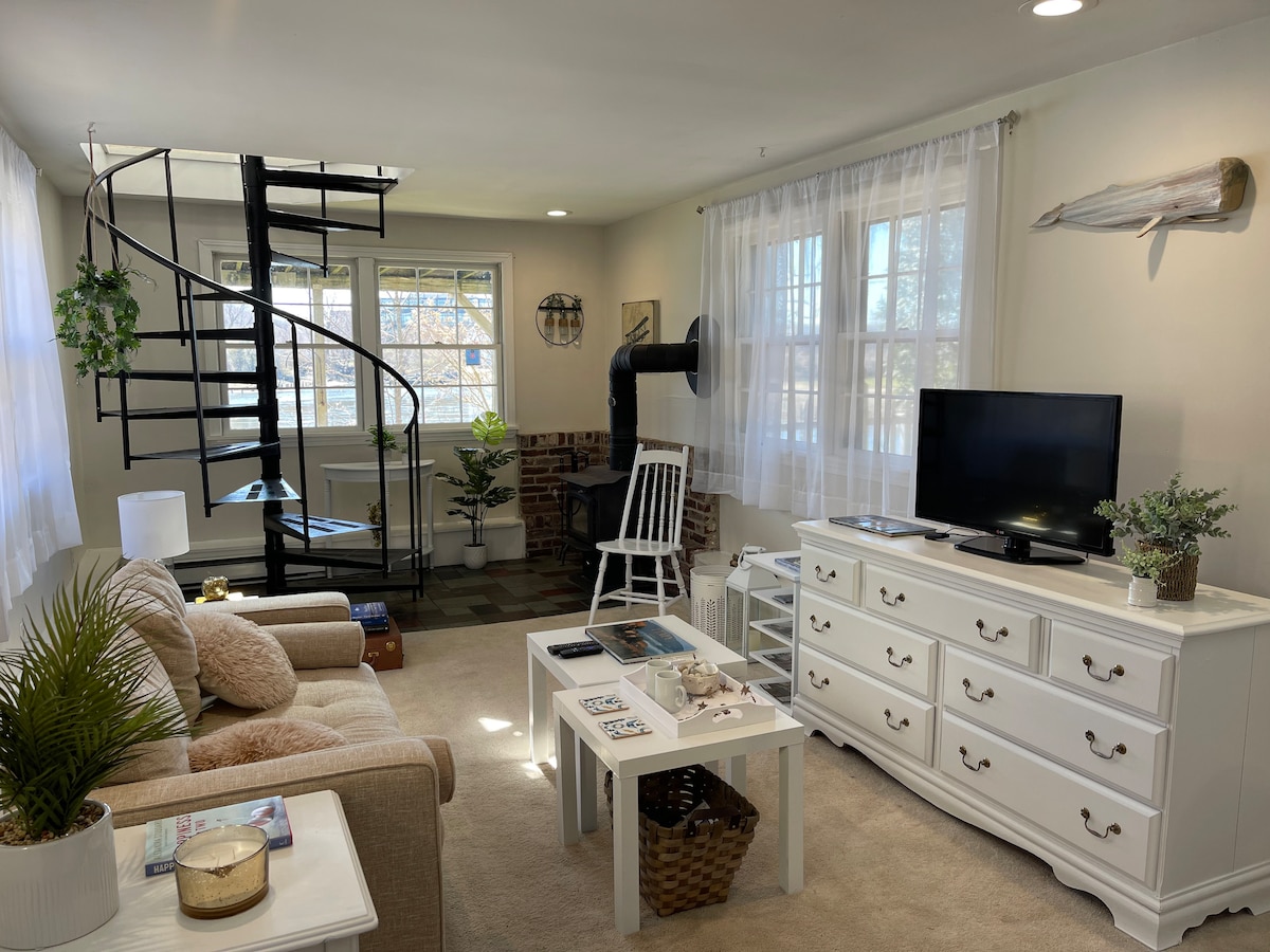 A light-filled living area features a spiral staircase ascending to the upper level. A cozy sofa is complemented by a white coffee table and a media console with a television. Large windows allow natural light to brighten the space, while indoor plants add a touch of nature.