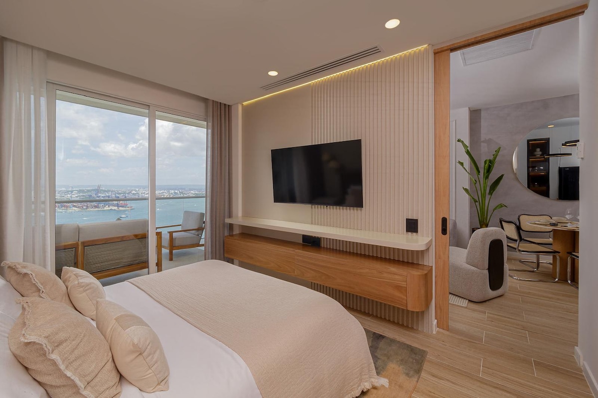 A modern bedroom features a spacious bed adorned with neutral-toned bedding. A wall-mounted television is positioned above a wooden console. Large windows offer views of the water and cityscape, while a cozy seating area and workspace are visible in the background.