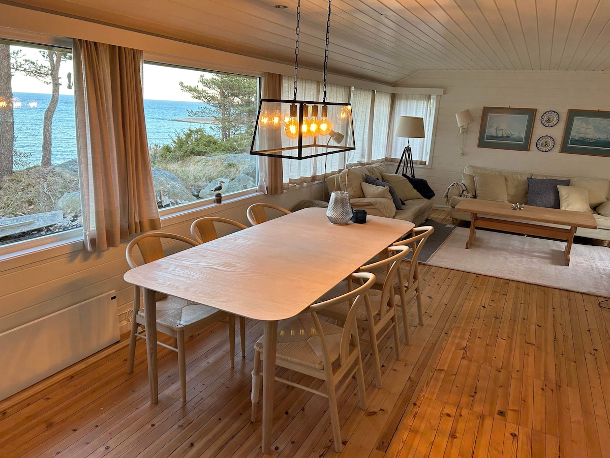 A spacious dining area is illuminated by a modern light fixture above a long wooden table, surrounded by six wooden chairs. Large windows offer views of the ocean and surrounding nature, while soft furnishings in neutral tones contribute to a calming ambiance.
