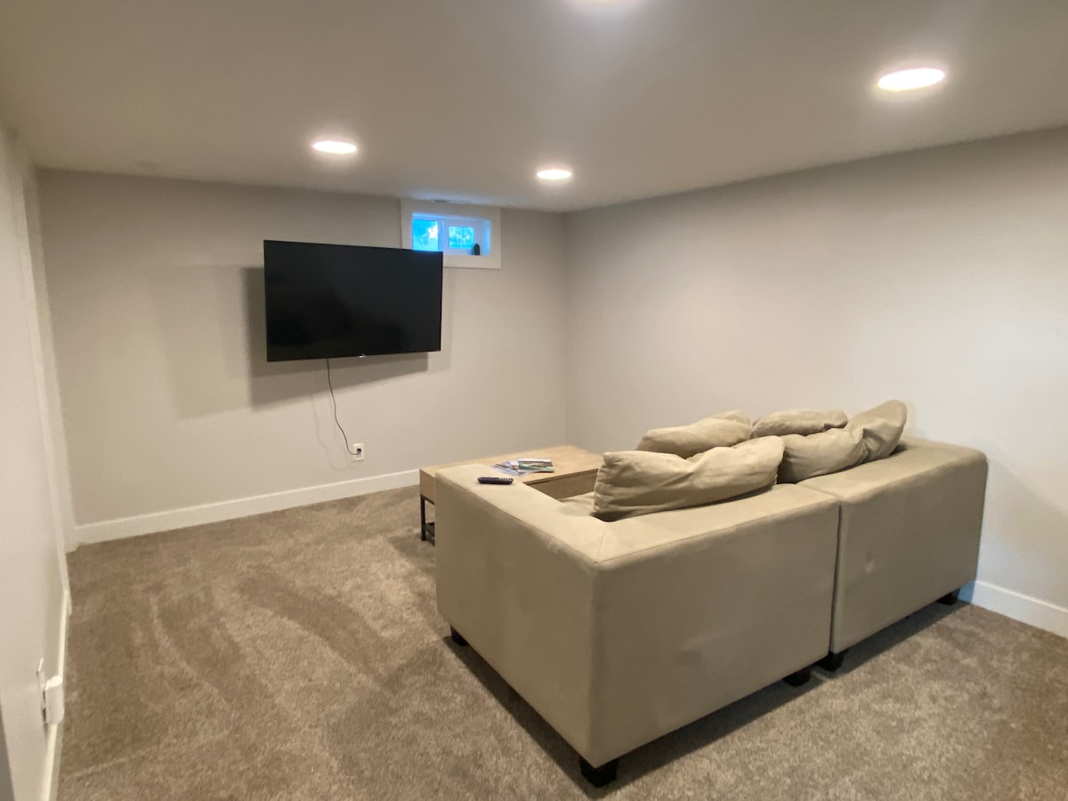 A cozy living room area is furnished with a plush, L-shaped couch facing a wall-mounted flat-screen television. Soft, neutral tones dominate the space, and natural light filters through a small window. The carpeted floor adds a comforting touch to the room.