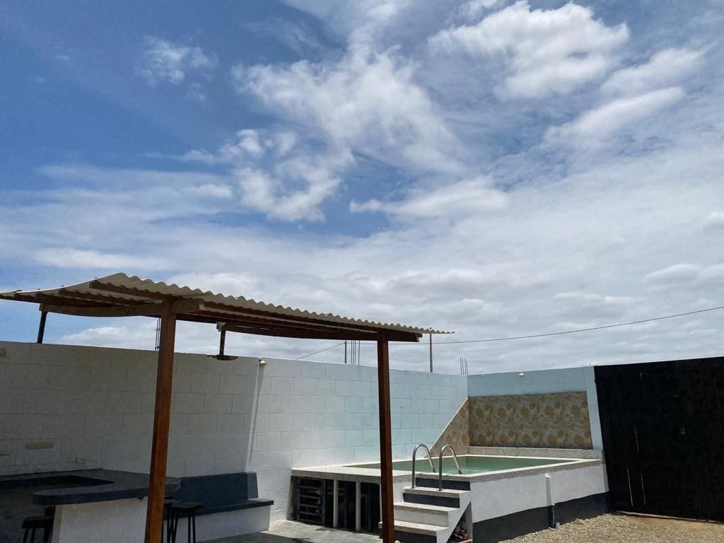 An outdoor area features a covered seating space with wooden beams and a table. A swimming pool is visible in the background, set against a light-colored wall, with the sky above dotted by clouds.