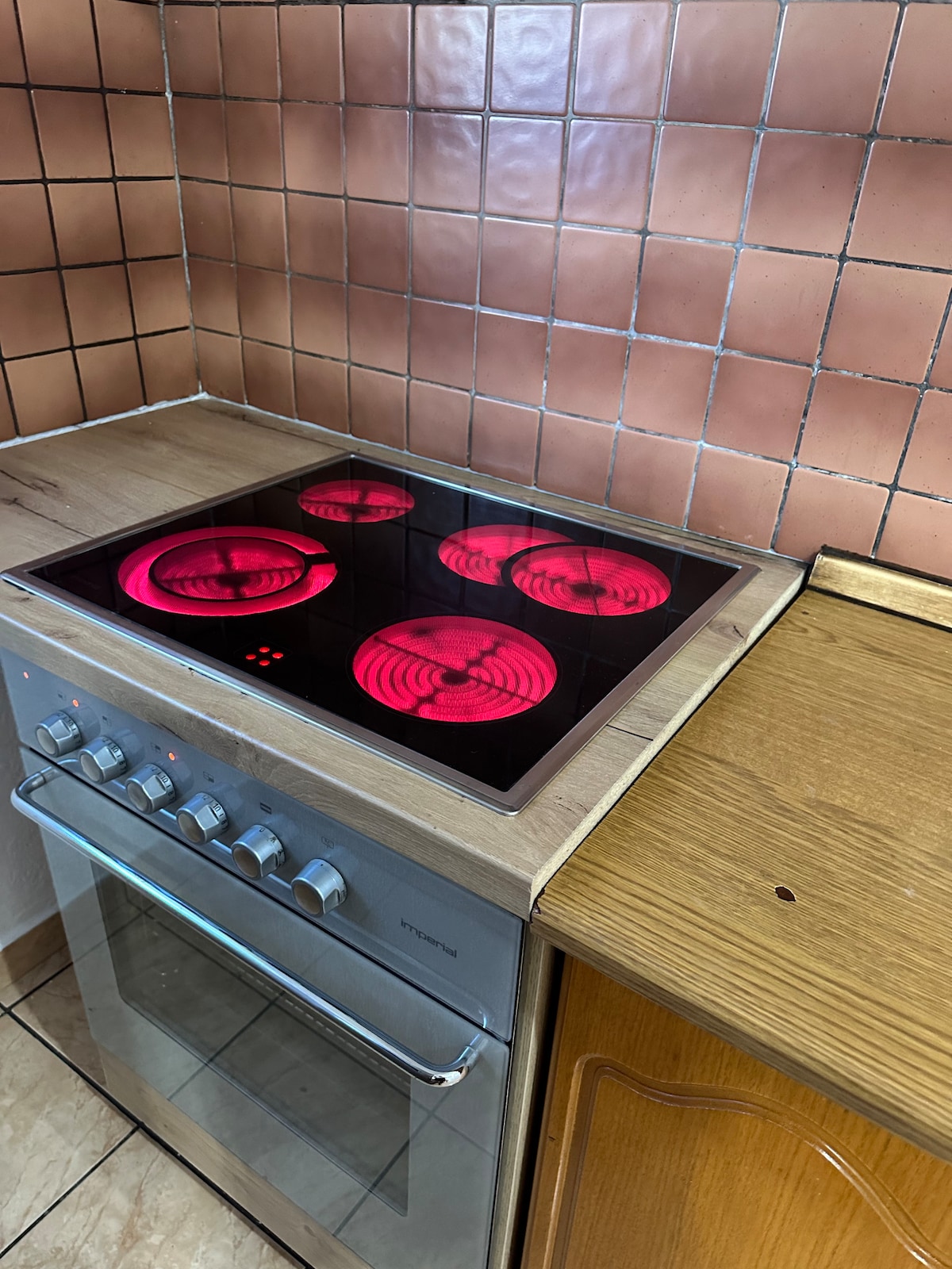 A modern cooktop features four illuminated burners against a backdrop of textured tiles. An oven is situated below, complemented by wooden cabinetry. The countertop is designed with a natural wood finish, highlighting the kitchen's functional layout.