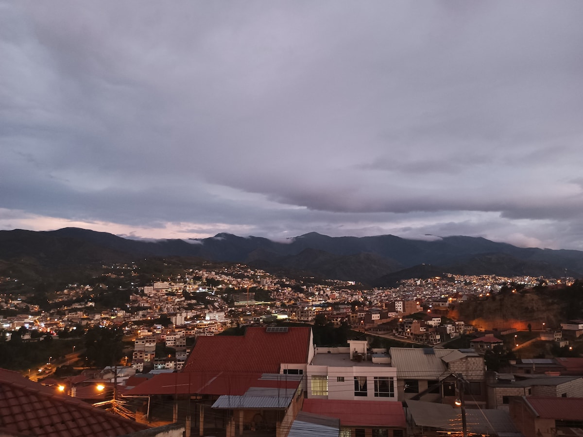 The image captures a panoramic view of the city illuminated at dusk, with a mixture of urban structures and natural landscapes. Mountains can be seen in the background, partially obscured by clouds, creating a serene atmosphere as the city lights begin to shine.