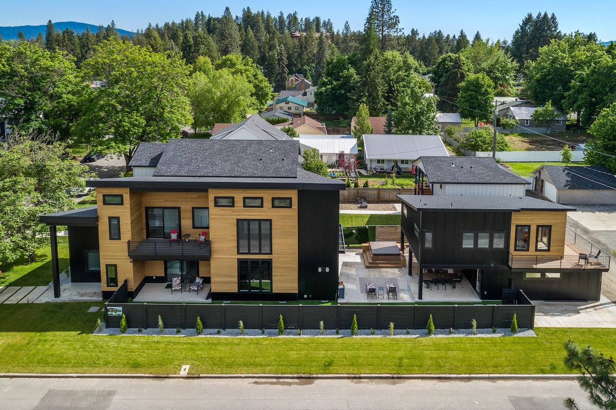 The exterior view showcases a modern guest house surrounded by landscaped grounds. Two distinct structures feature a mix of wood and dark siding. Spacious outdoor areas are visible, with designated seating and a patio space beneath a sun awning. Lush greenery frames the property.