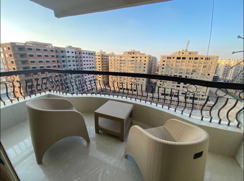 A balcony is furnished with two wicker chairs and a small wooden table, offering space for relaxation. The view showcases a cityscape with various buildings under a clear sky, allowing natural light to illuminate the area.