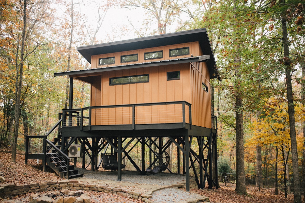 A modern treehouse is elevated on stilts, showcasing a wooden exterior with large windows. The surrounding area features autumn foliage, creating a serene atmosphere. A set of stairs leads to a spacious deck, complementing the structure's design.