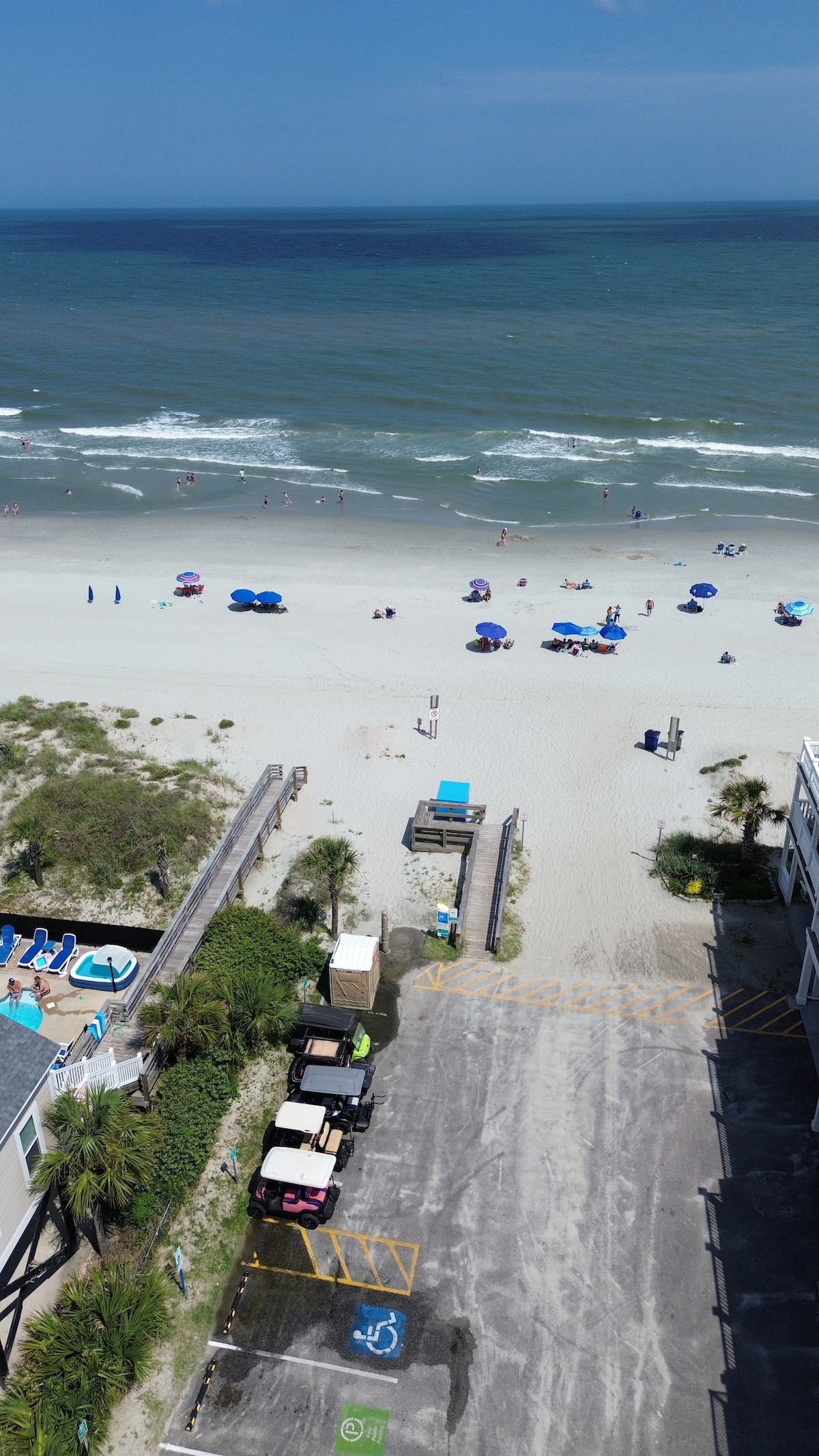 A view captures the nearby beach, showcasing soft sandy shorelines with beachgoers under large umbrellas. The ocean waves create gentle patterns along the shore. A parking area and pool are visible in the foreground, enhancing the accessibility to the beach.
