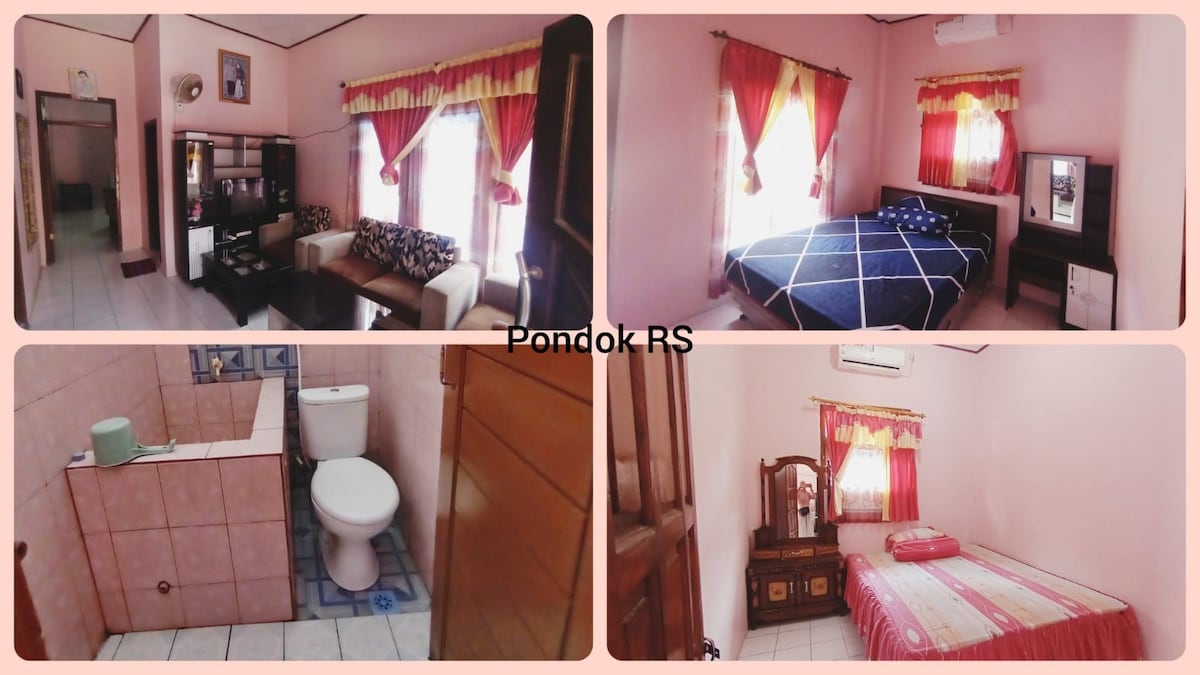 A welcoming room with soft pink walls features a spacious bed draped in a blue quilt. A modern dresser and a comfortable seating area are present. Bright curtains allow natural light to illuminate the space. A bathroom area is partially visible, equipped with essential amenities.