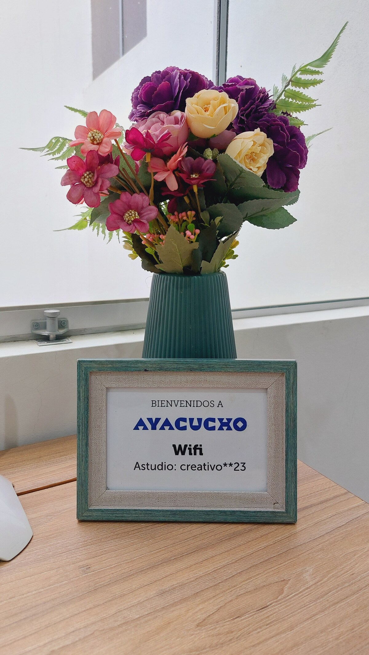 A decorative arrangement of colorful artificial flowers is displayed in a green vase on a wooden table. A welcome sign is framed, featuring the text 'BIENVENIDOS A AYACUCHO' and 'Wifi' prominently, creating an inviting atmosphere for guests.