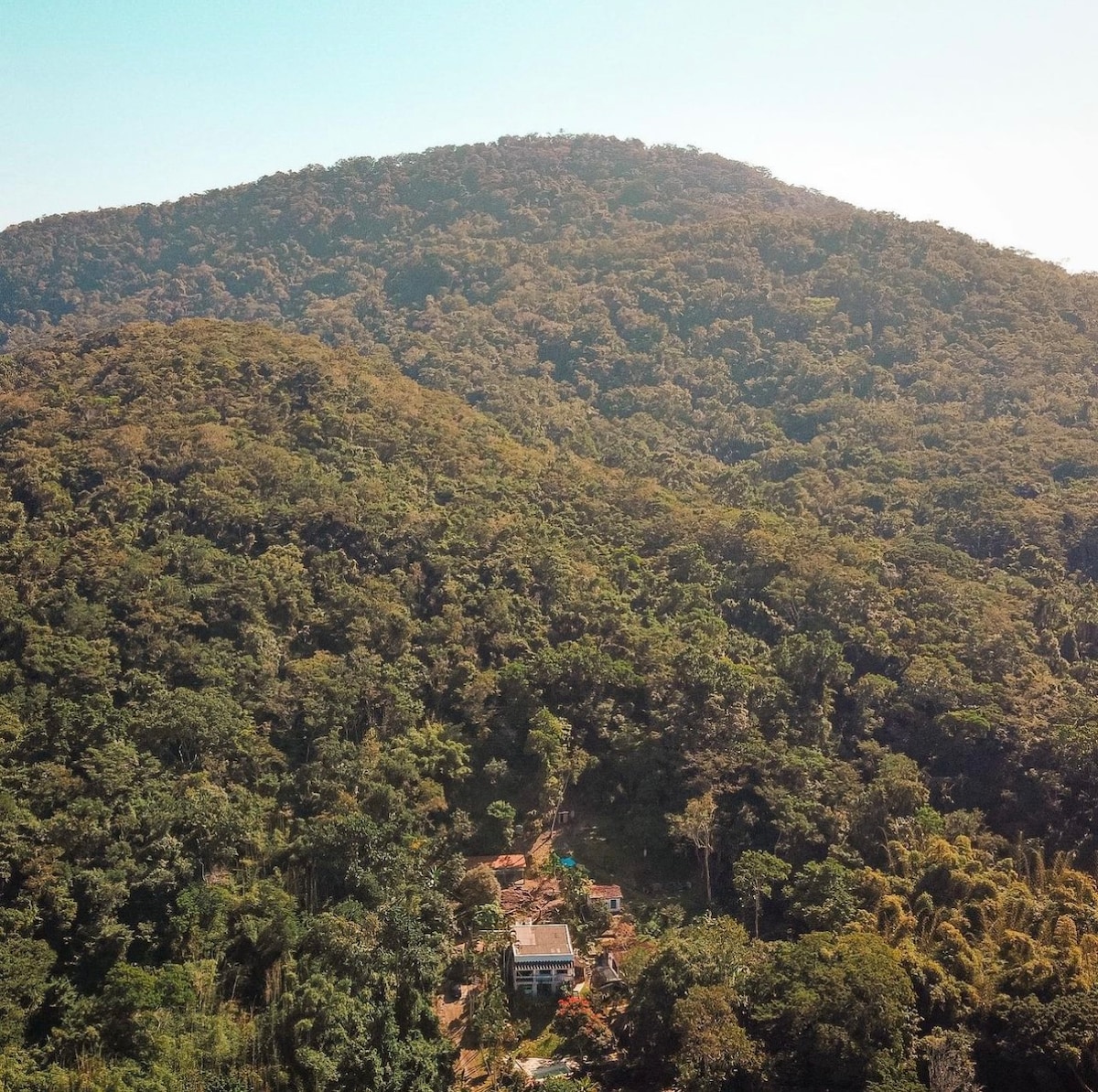An aerial view captures a secluded property nestled within dense greenery at the base of a mountain. Lush trees surround the area, providing a sense of privacy and tranquility, while the mountainous backdrop enhances the natural beauty of the location.
