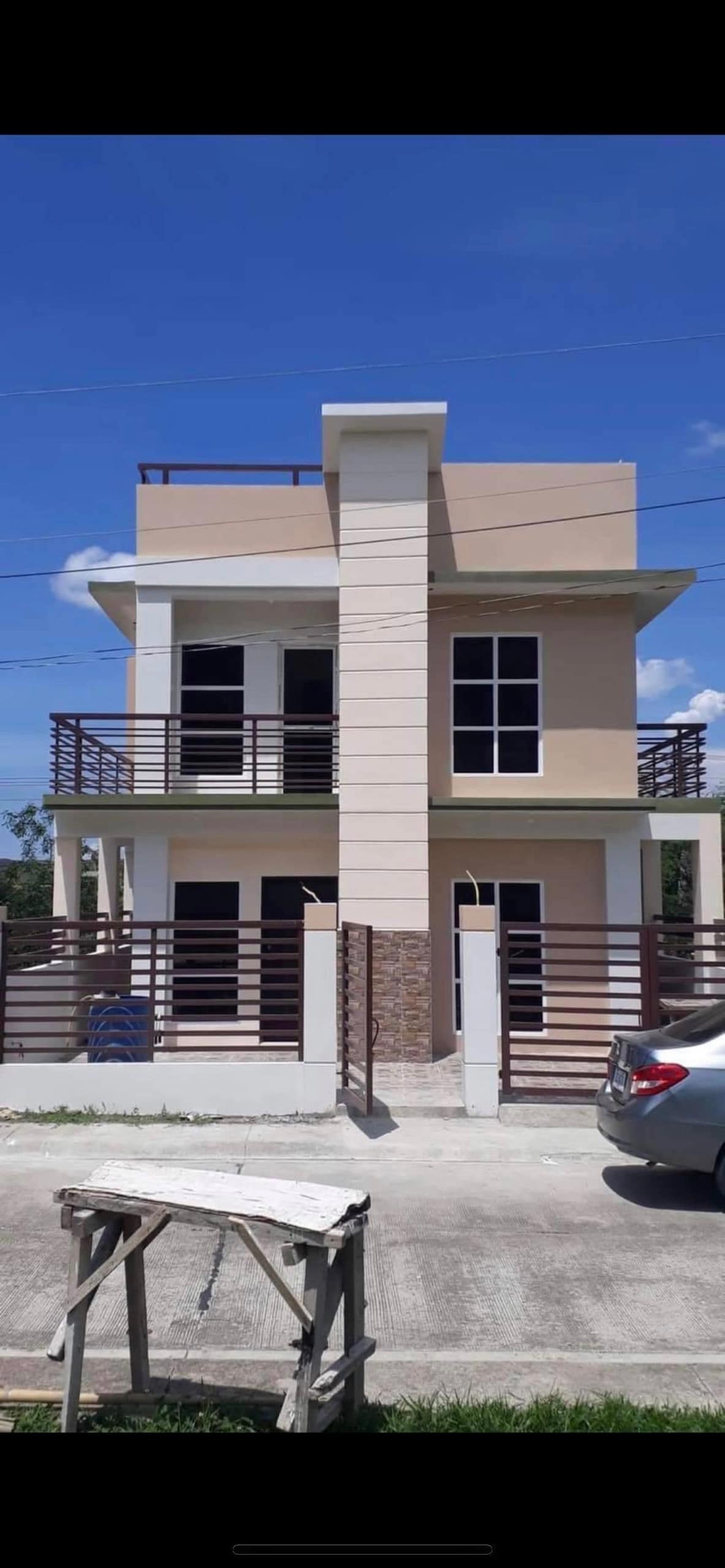 The exterior of a two-story house is displayed, featuring a light-colored facade with multiple windows and balconies. A car is parked in the foreground, and the driveway is clearly visible. The sky above is bright with a few scattered clouds.