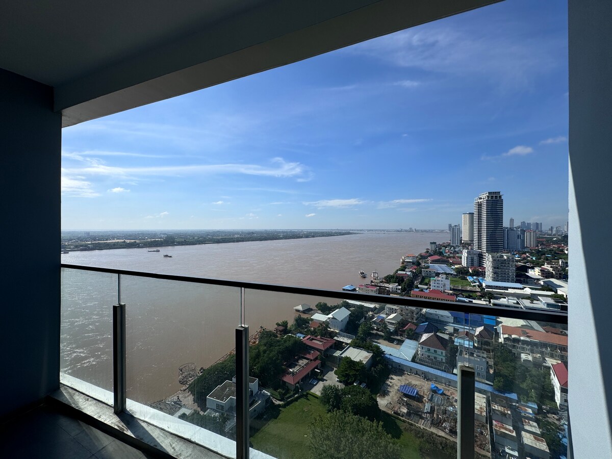 A panoramic view of the Mekong River is framed by a large window, highlighting the river's brown waters and distant cityscape. Buildings and residential areas are visible along the riverbanks, under a clear blue sky with scattered clouds.