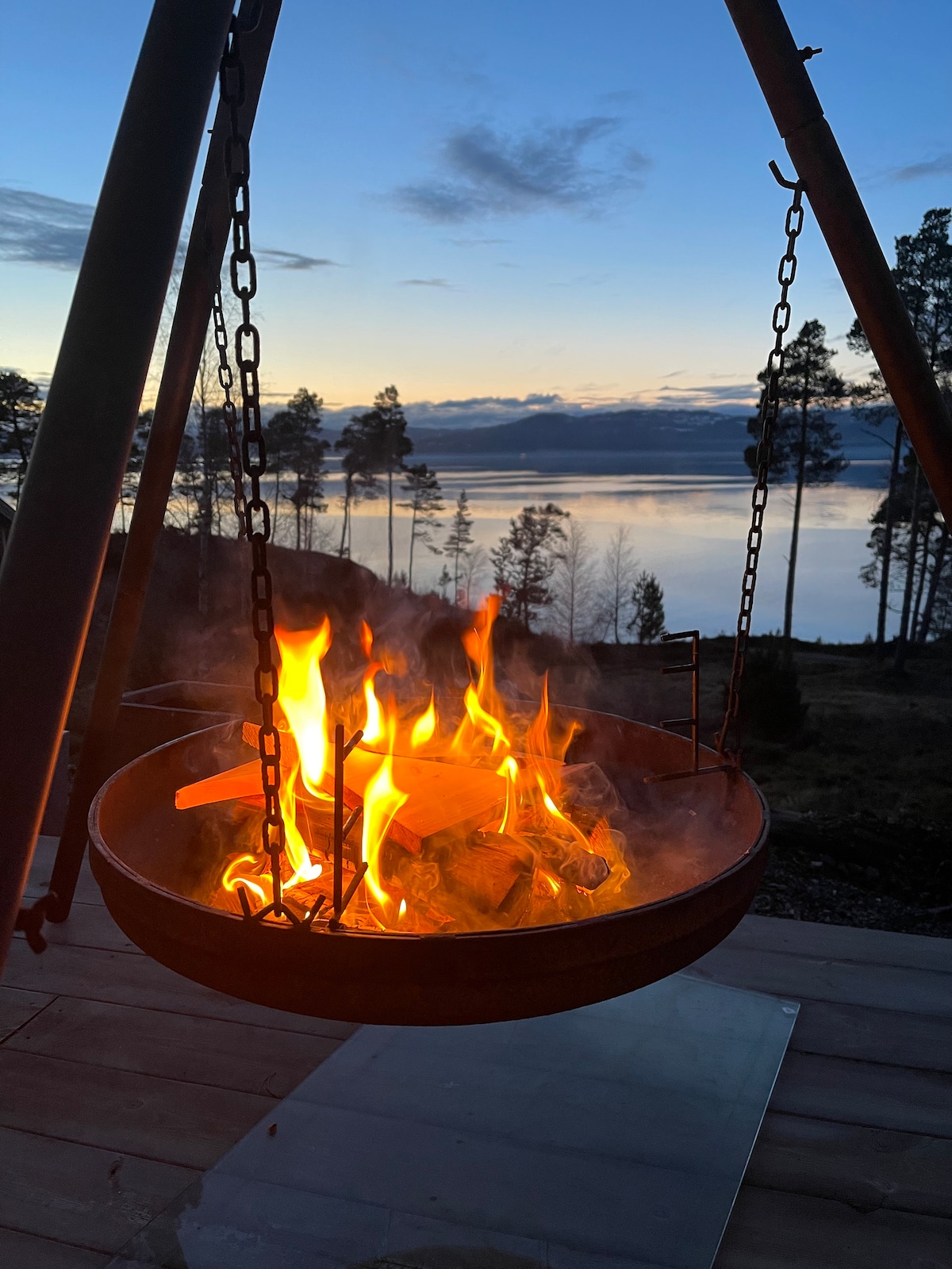 A fire pit is suspended from a frame, with flames dancing above a circular grill. The backdrop features a serene lake reflecting the colors of the early evening sky, framed by trees. Smoke gently drifts upwards as the sun sets, casting a peaceful ambiance.
