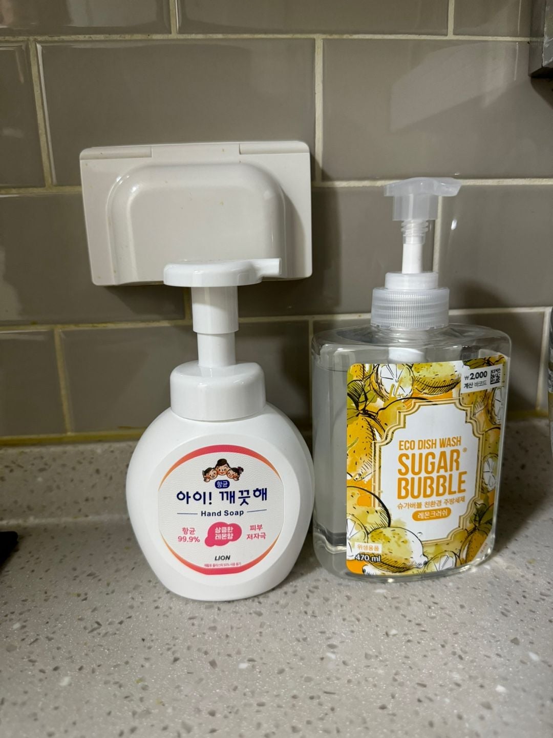 A sink area displays two soap dispensers on a countertop. On the left, a white pump bottle labeled as hand soap features a cartoon design. To the right, a transparent pump bottle contains eco dish wash, adorned with a lemon-themed label.