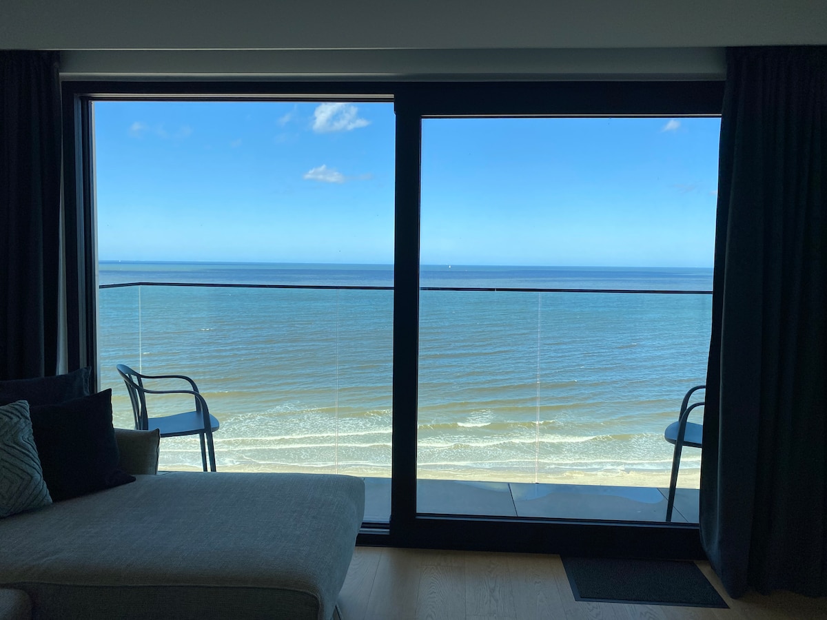 A modern living area features large glass doors offering unobstructed views of the sea. Two chairs are placed on the balcony, allowing for outdoor seating. The calm blue sky complements the shimmering water, creating a serene coastal atmosphere.