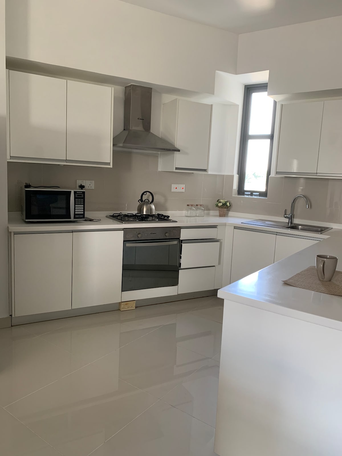 A modern kitchen is equipped with sleek white cabinetry and stainless steel appliances. A gas stove is prominently featured alongside a microwave and a kettle. Natural light enters through a window above the sink, illuminating the clean countertops and tiled flooring.