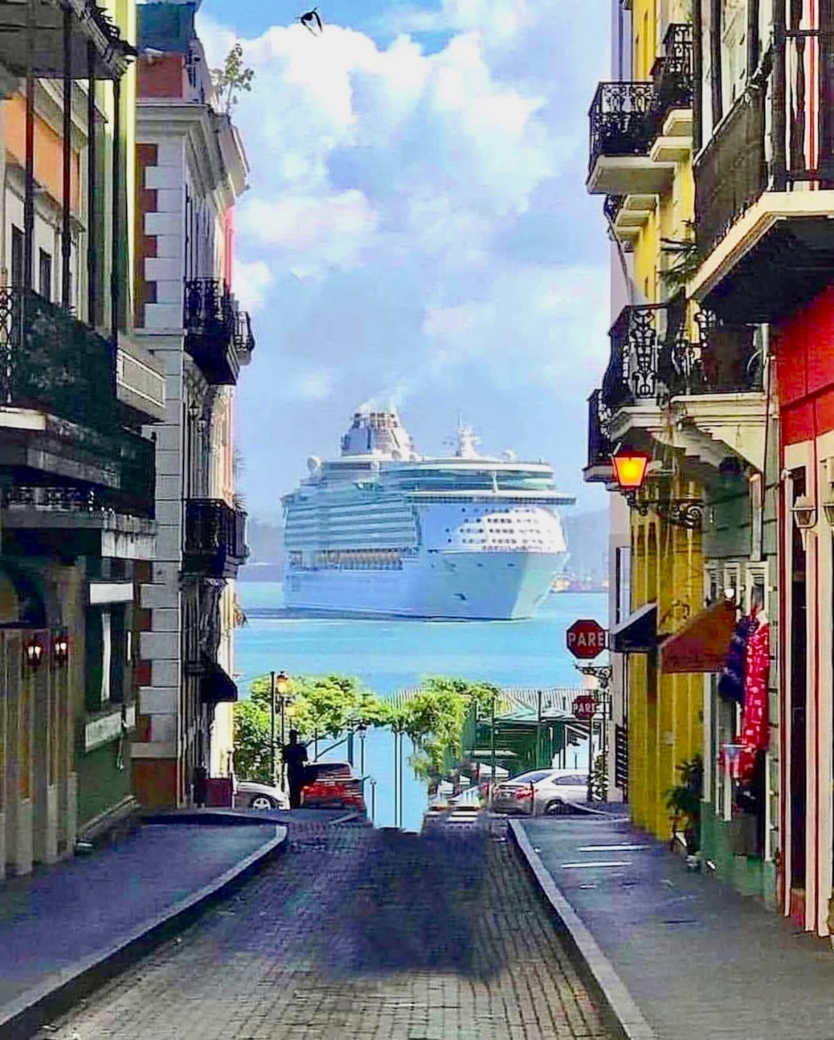 A street view captures a cruise ship anchored in the distance, framed by colorful buildings with intricate balconies. Sunlight reflects off the water, with palm trees and traffic signs visible along the curved road, offering a glimpse of the vibrant urban landscape.