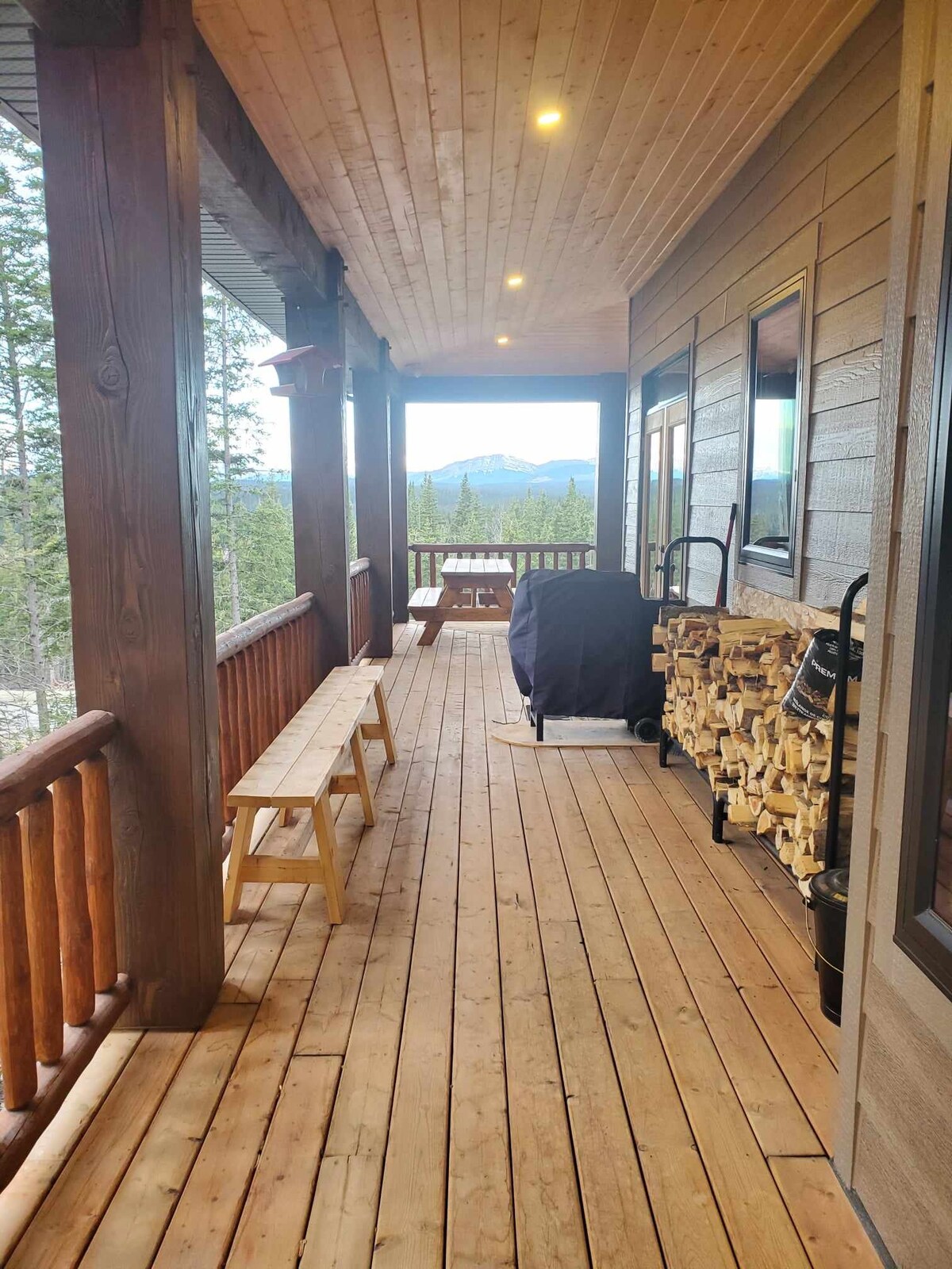A covered wrap-around deck features wooden benches along the side, with neatly stacked firewood on display. A gas grill is visible, set against a backdrop of towering trees and distant mountain peaks, creating a serene outdoor space for relaxation.