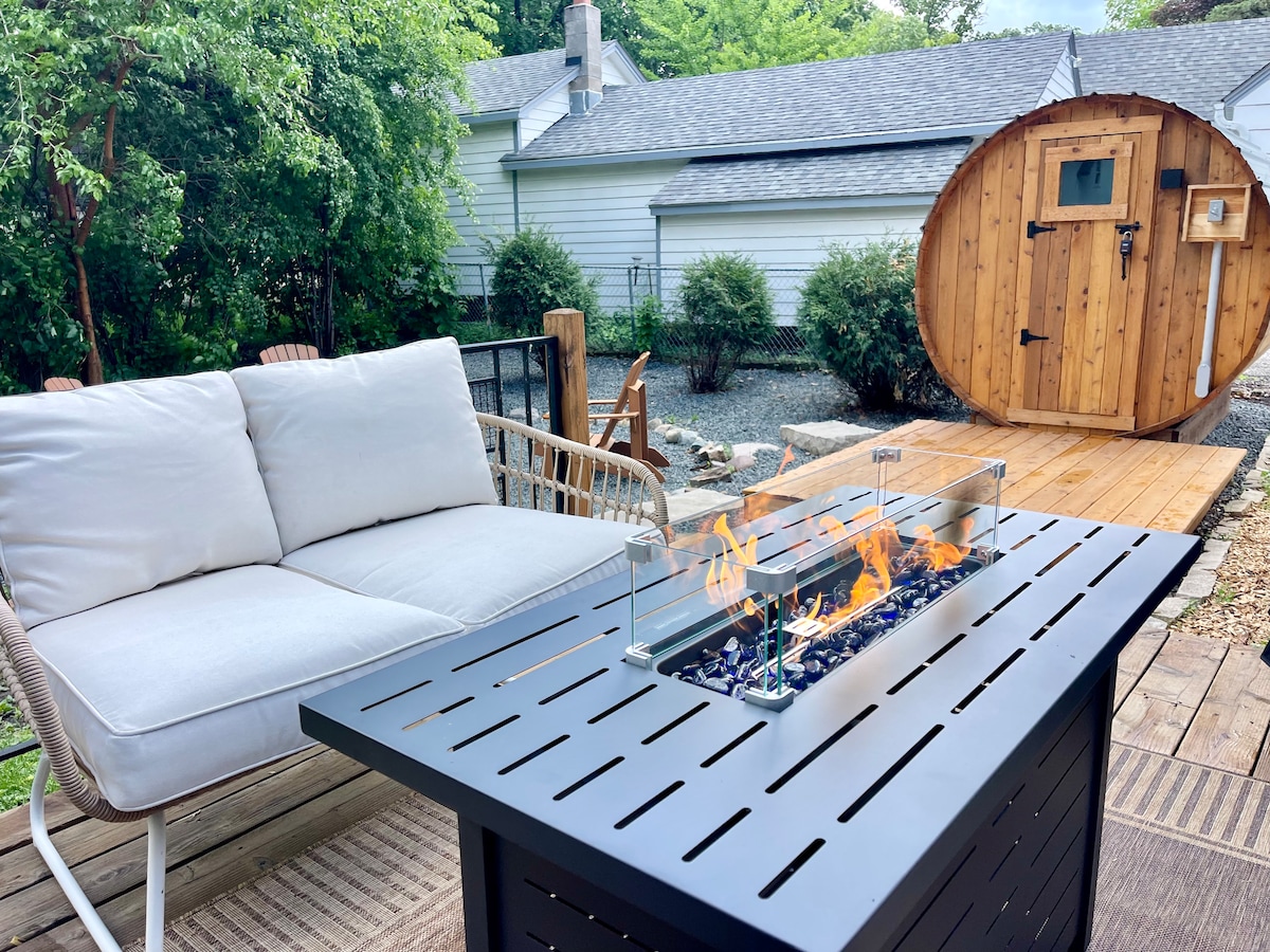 An outdoor seating area is highlighted by a modern fire pit with a glass barrier, featuring vibrant flames. A cozy sectional sofa offers comfortable seating, set against a backdrop of greenery and a uniquely designed barrel sauna.