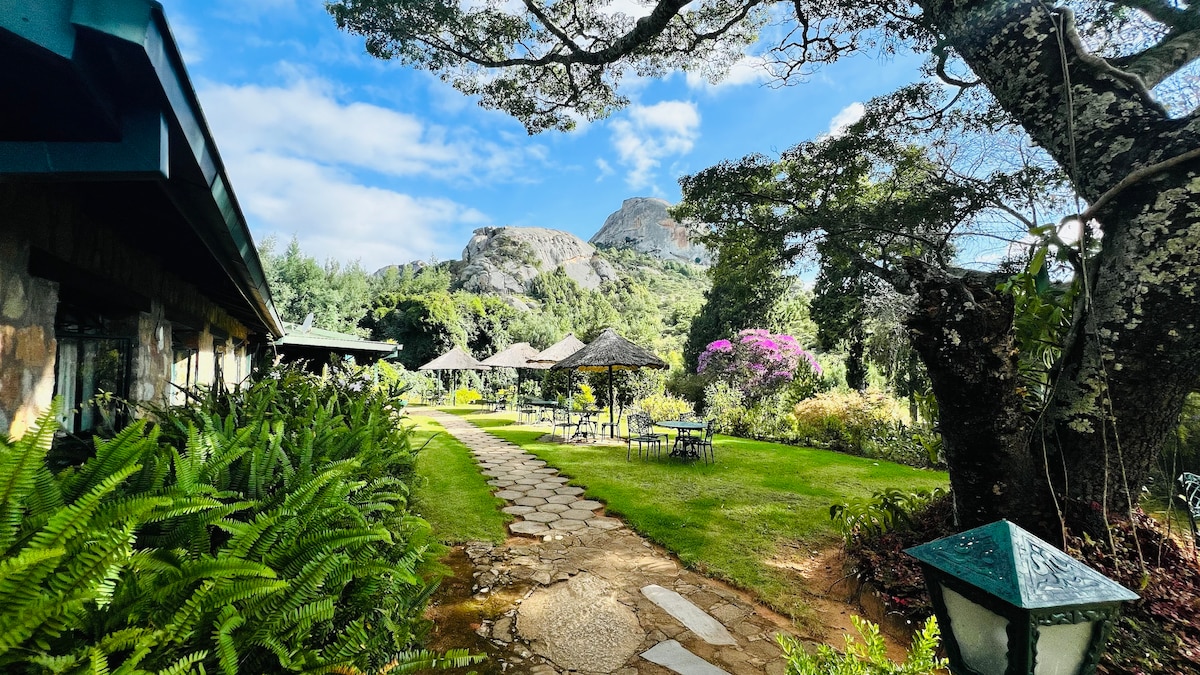 A stone pathway gently winds through well-maintained gardens, flanked by lush greenery and flowering plants. Scenic mountain views can be seen in the background, while shaded seating areas with thatched umbrellas are positioned along the pathway, inviting relaxation amidst nature.