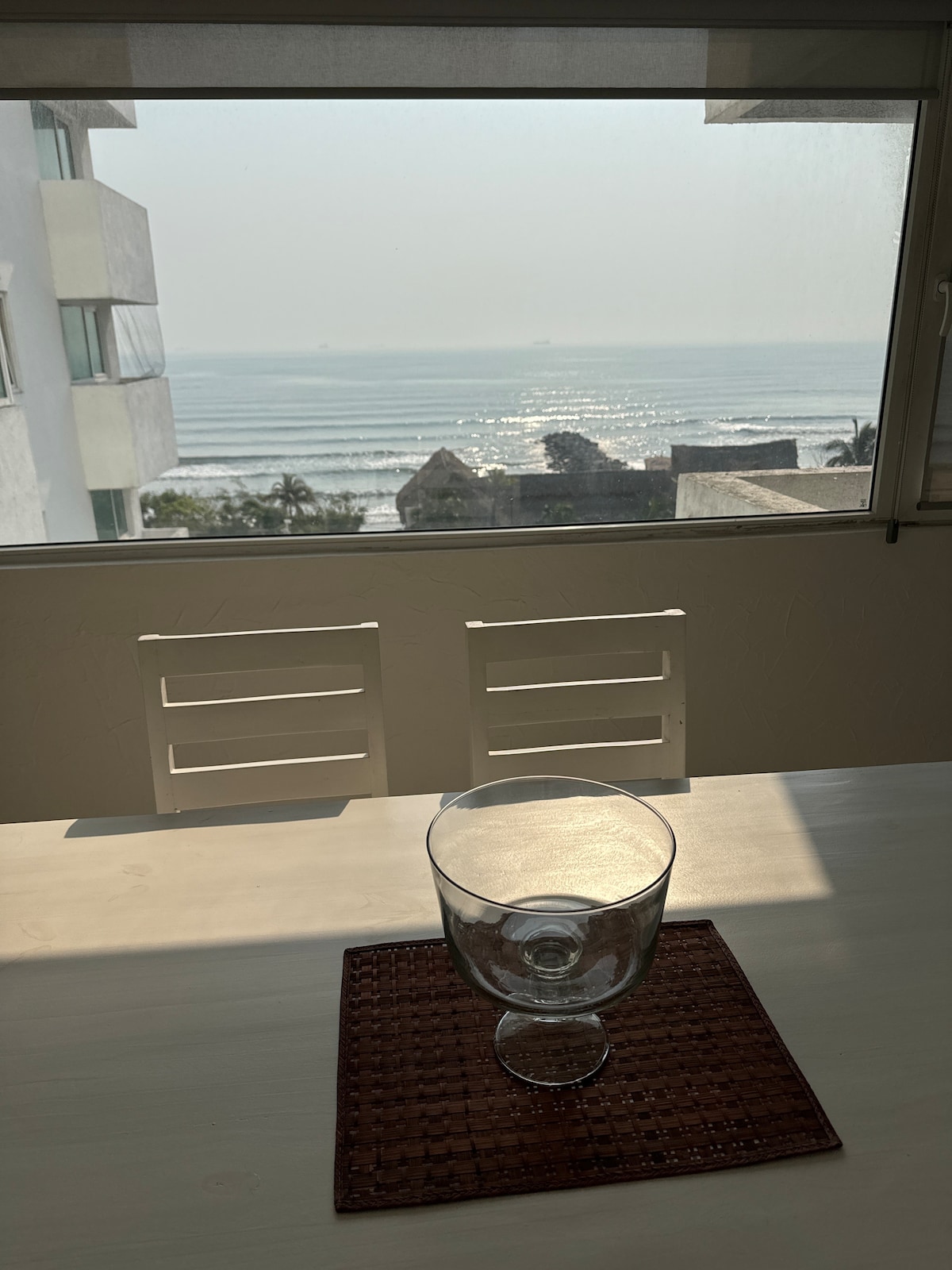 A dining table is set beside a large window showcasing a view of the ocean. The sunlight reflects off the water, creating a serene atmosphere. A clear glass bowl sits on a woven placemat, complementing the minimalist decor.