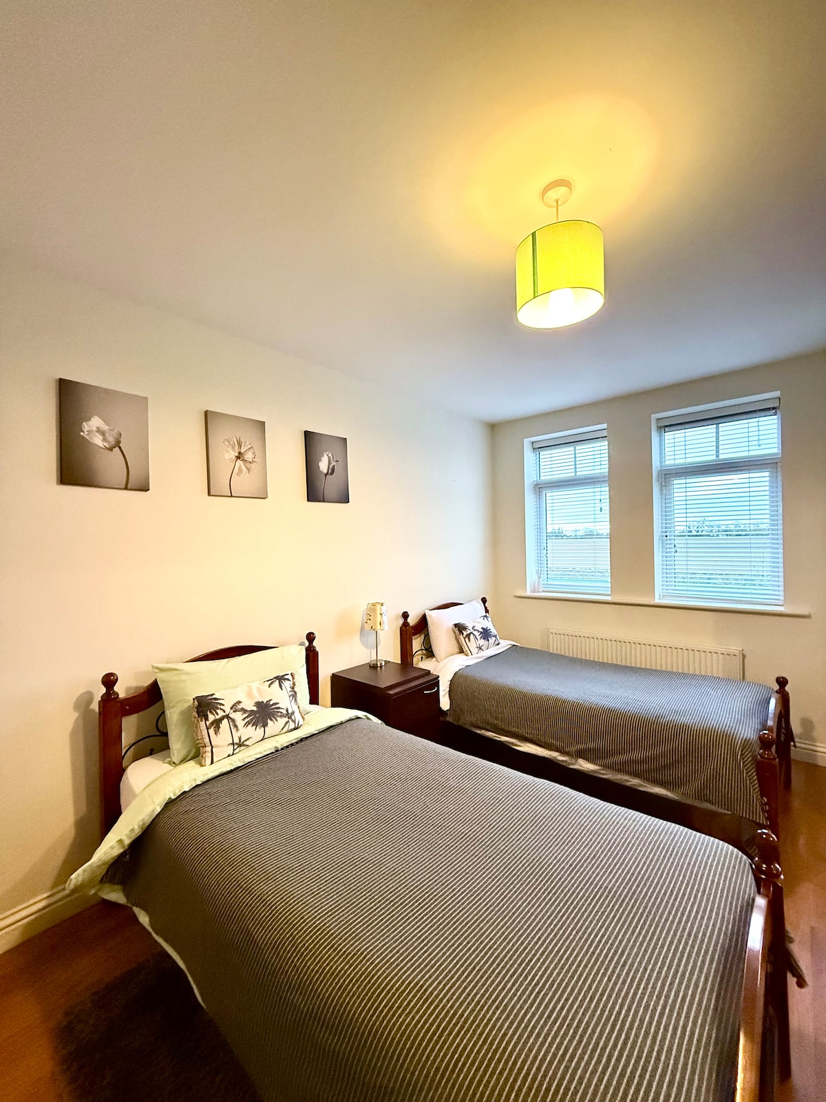 A light-filled private room features two single beds with neutral bedding. Decorative artwork is displayed on the wall above the beds. Natural light enters through two large windows with blinds, illuminating the warm wooden floor.