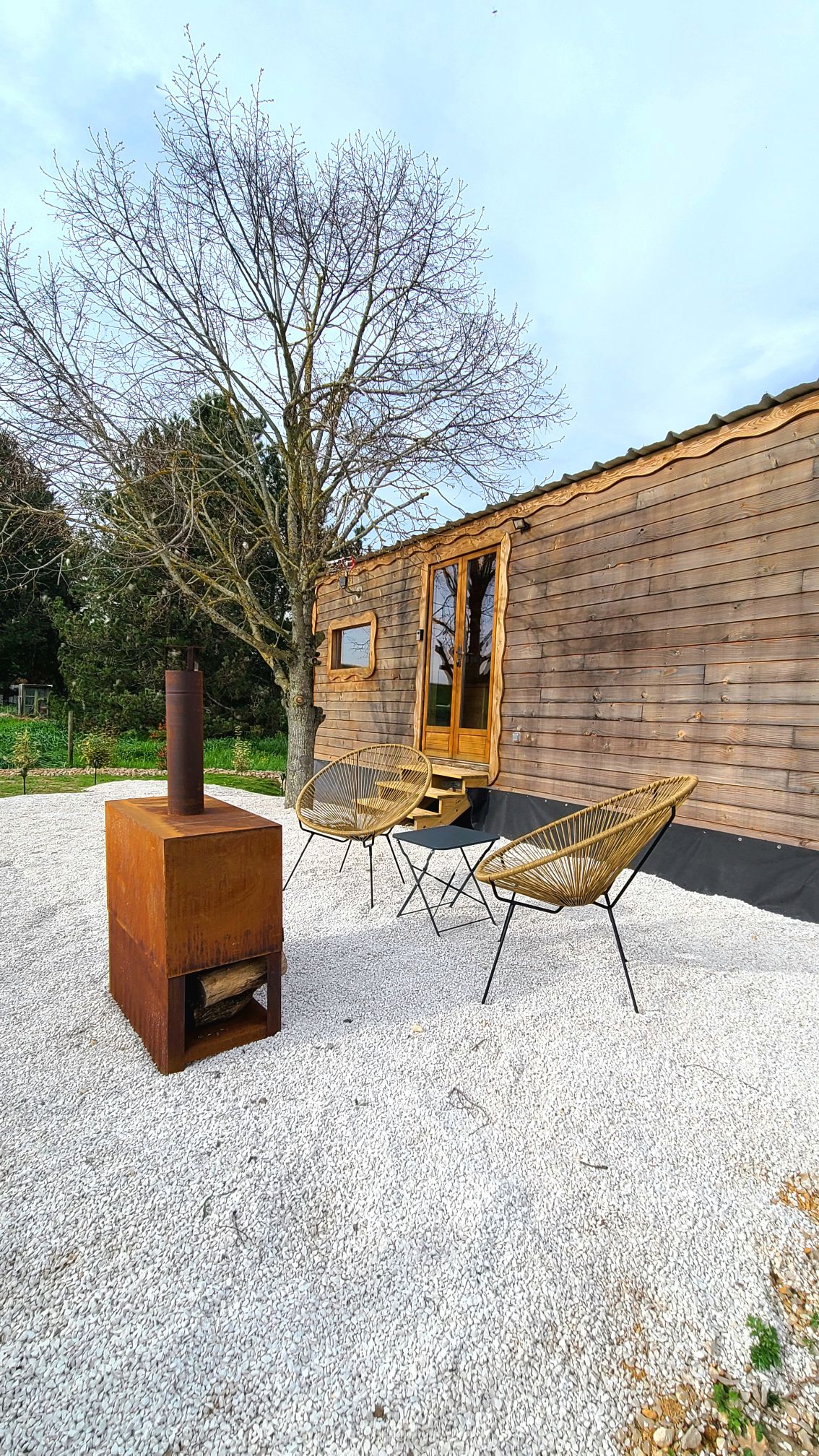 An outdoor seating area is visible, featuring two woven chairs positioned near a small metal fireplace. The wooden exterior of the Tiny House is seen in the background, complemented by a tree and a gravel surface in the foreground.