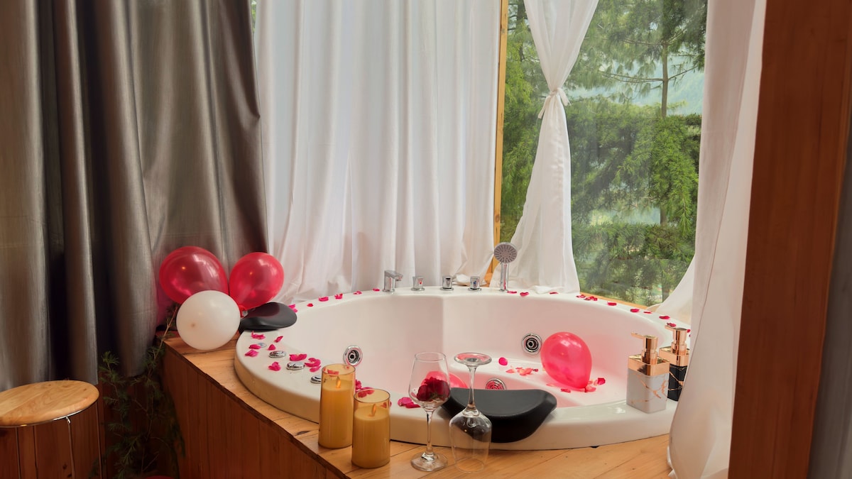 A luxurious jacuzzi tub is positioned in a corner, surrounded by smooth wooden panels. The area is decorated with rose petals, balloons, and lit candles. Sheer curtains allow soft light to filter through, offering views of the serene outdoor landscape.