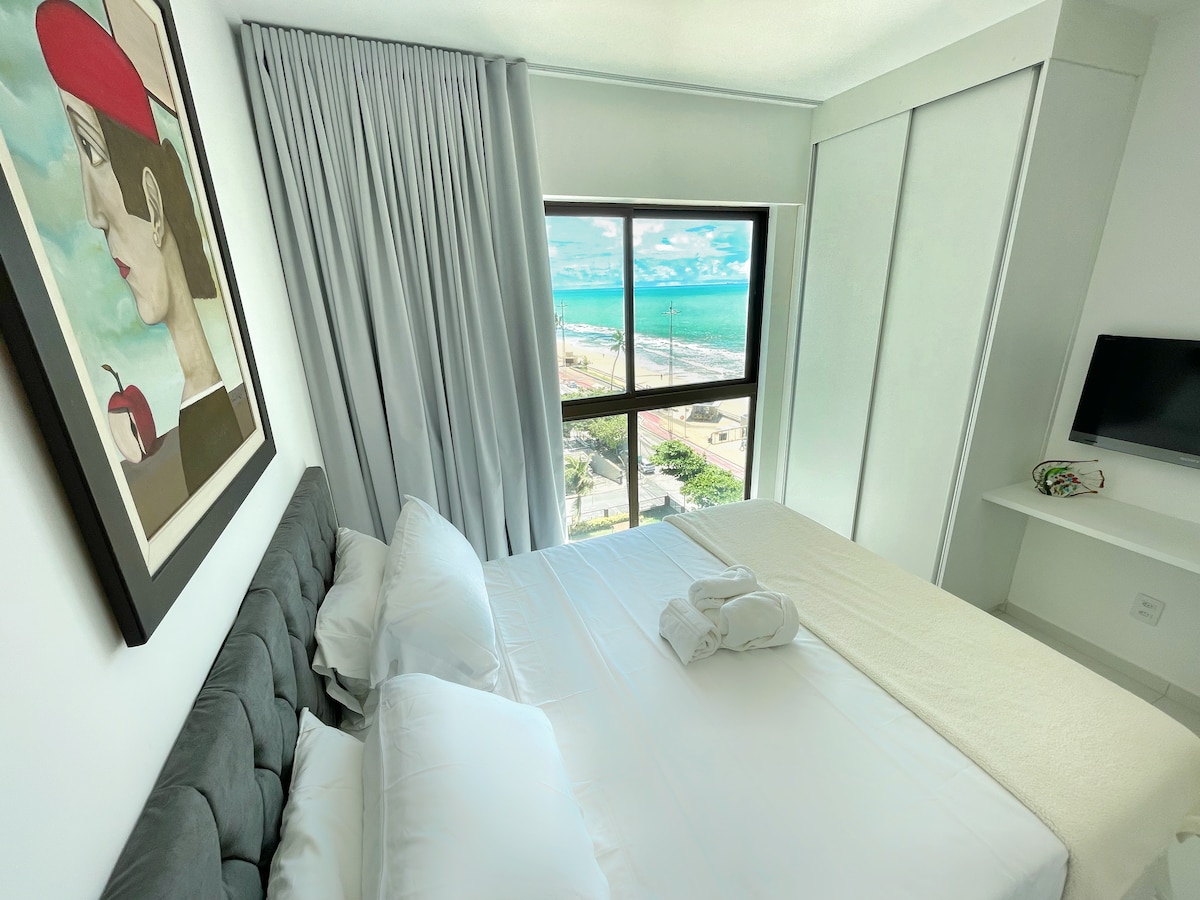 A serene bedroom features a spacious bed with light bedding. Large windows provide a view of the ocean and surrounding area. Soft gray curtains frame the windows, and a piece of artwork is displayed on the wall, adding an artistic touch to the space.