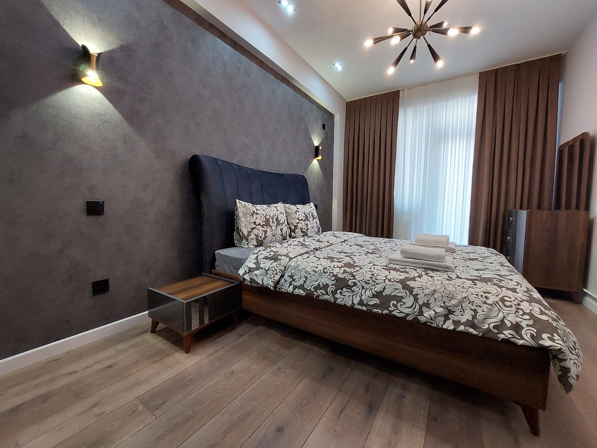 A comfortable bedroom features a large double bed adorned with patterned bedding. Soft lighting illuminates the space, with wall-mounted fixtures creating a cozy atmosphere. Natural light filters through sheer curtains, and a wooden bedside table complements the hardwood floor.
