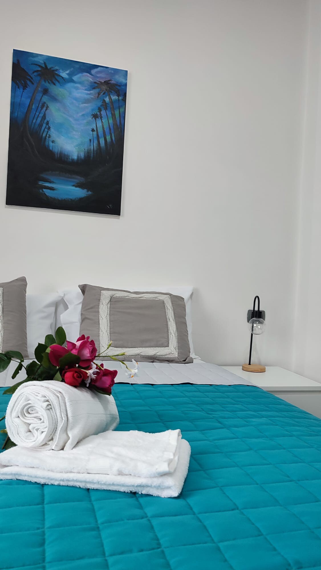 A neatly made bed is complemented by gray pillows and a teal quilt. A small vase with flowers is placed near a stack of rolled towels. A framed painting depicting a serene landscape hangs on the wall above the bed.
