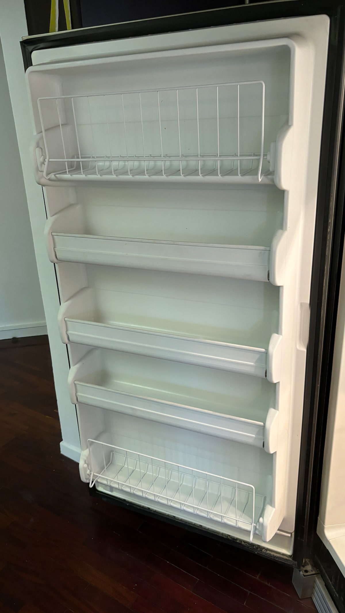 A refrigerator door is shown, featuring multiple shelves and compartments designed for storage. The interior is illuminated, highlighting the white shelving and clean lines. The sleek exterior contrasts with the wooden floor, emphasizing the refrigerator's modern design.