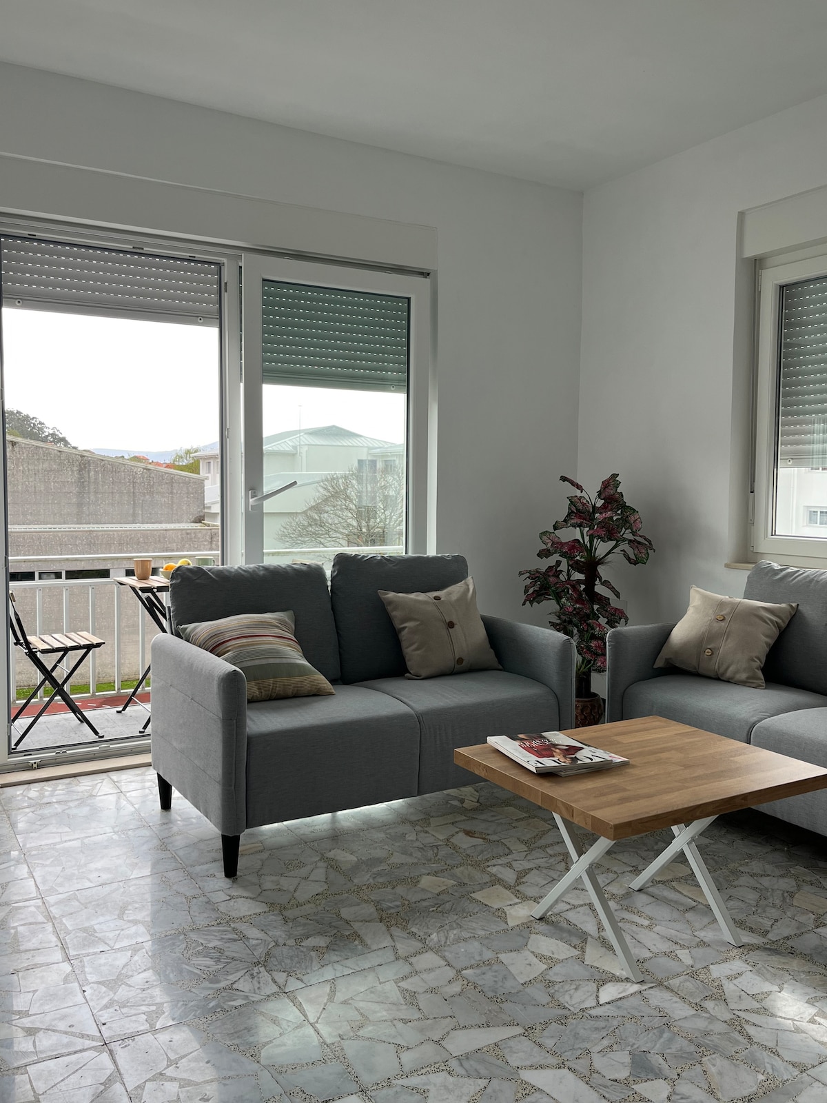 A bright living area features a pair of gray sofas adorned with decorative cushions. A wooden coffee table rests in front, magazines neatly arranged on top. Large sliding doors open onto a small terrace, allowing natural light to fill the space.