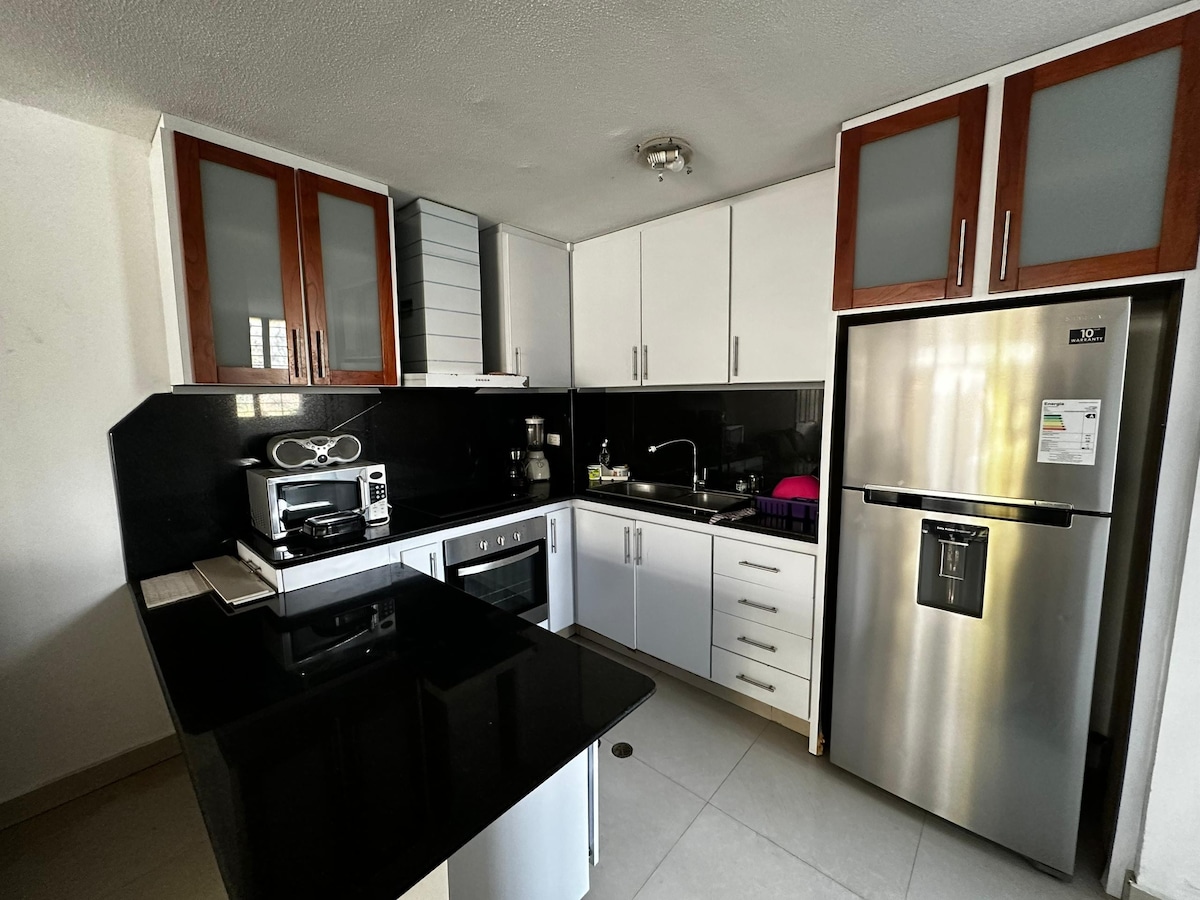 The kitchen features modern cabinetry with a mix of wood and white finishes, complemented by a dark countertop. Essential appliances include a microwave, toaster, and refrigerator. Natural light enters through a nearby window, illuminating the clean and organized workspace.