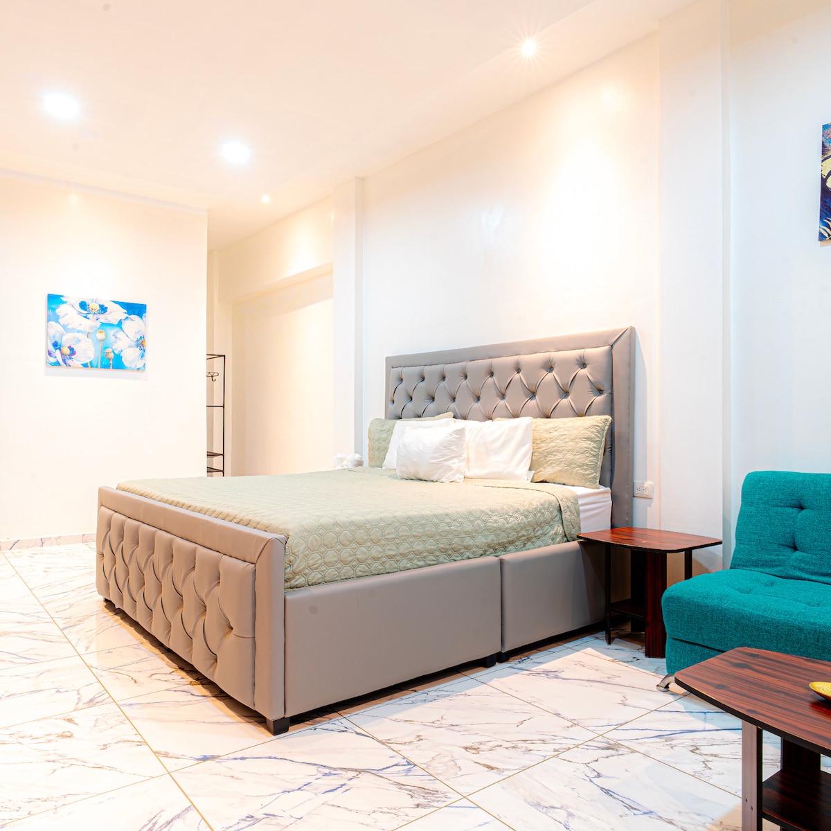 A spacious sleeping area features a comfortable bed with a tufted headboard, accompanied by a vibrant turquoise chair. Soft lighting illuminates the room, highlighting the marble flooring and decorative wall art.